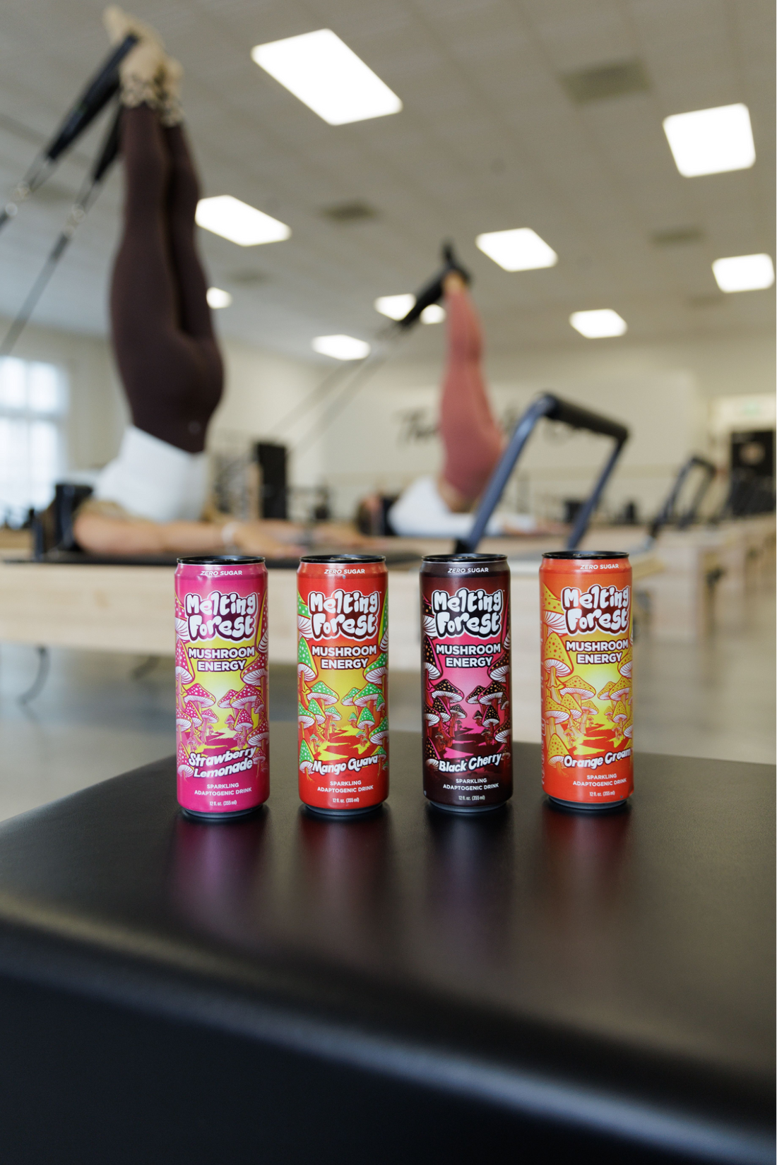 Four mushroom drinks in line on a black surface. In the background, there are women doing pilates